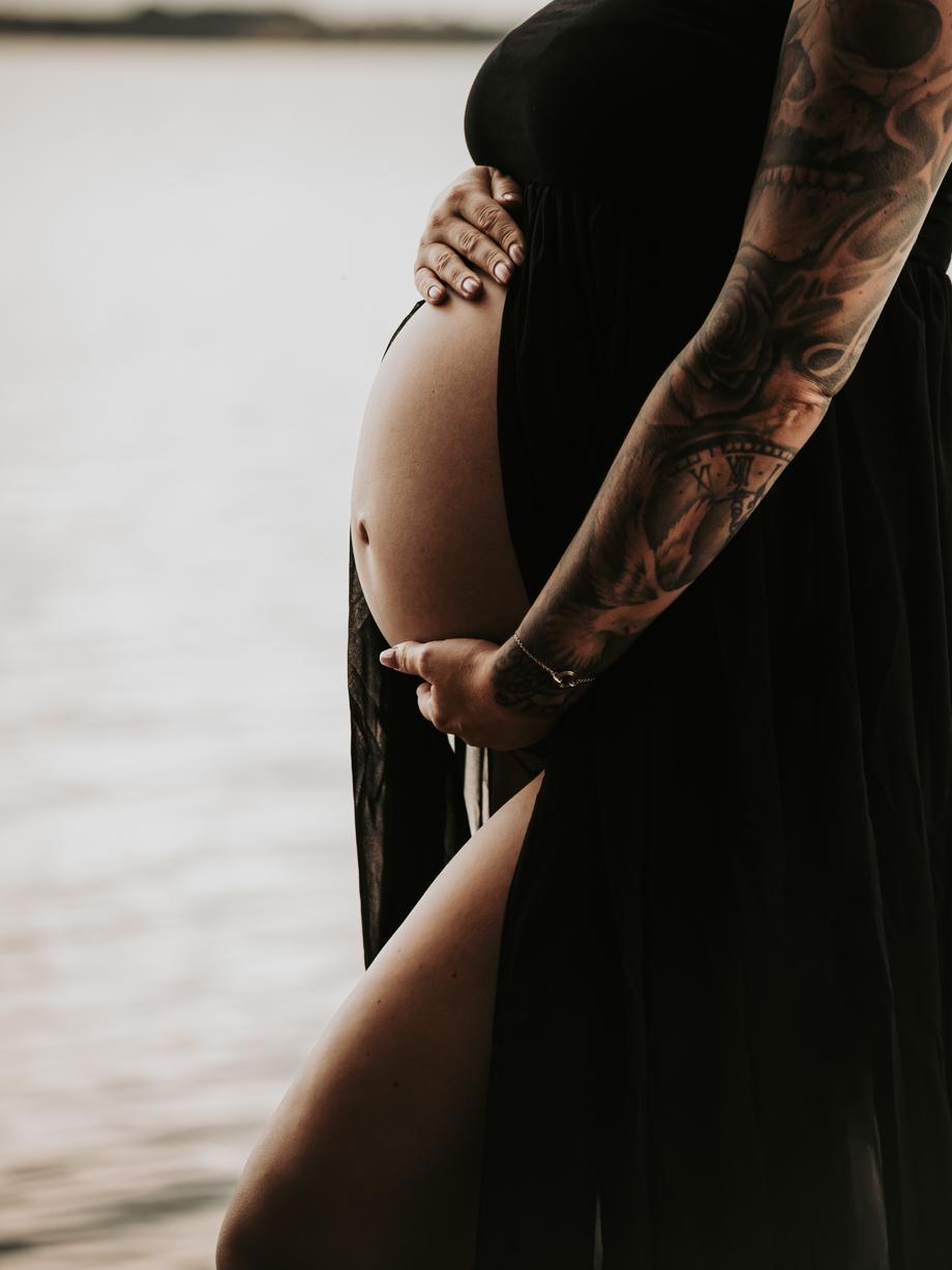 Schwangere im schwarzen Kleid hält ihren Babybauch, im Hintergrund das Wasser